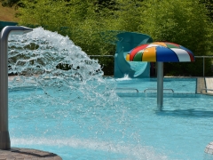Spaßbecken mit einem Wasserspeier links, der Wasserrutsche im Hintergrund und einem Wasserpilz rechts