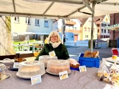 Eine Verkäuferin steht hinter ihrem Verkaufsstand. Im Vordergrund stehen Kuchen