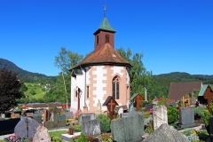 Im Vordergrund Grabsteine von hinten, dahinter die Friedhofskapelle. Weiß verputzt, Fenster und Ecken mit Sandstein