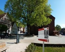 Rechts vorne Museums-Schild, im Hintergrund katholische Kirche und großer Lindenbaum