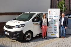 Im Hintergrund ist ein weißer Kleinbus mit Werbeaufdrucken zu sehen. Davor stehen Sarah Huber und Bürgermeister Uwe Gaiser, zwischen Ihnen ein Roll-Up mit allen Sponsorenlogos